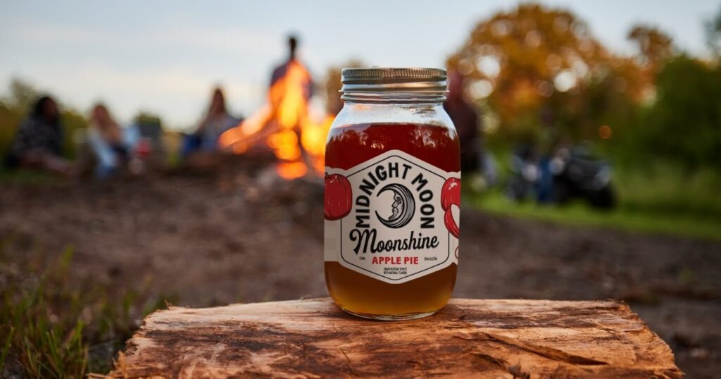 Midnight Moon Apple Pie Moonshine on log next to campfire. 