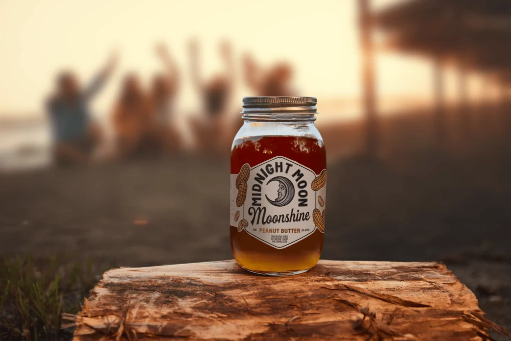 An opened jar of Midnight Moon Peanut Butter Moonshine on the beach at sunset.